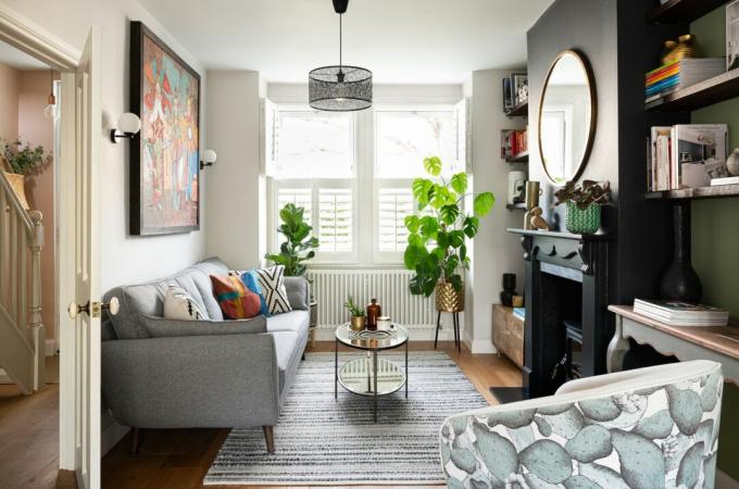 Stue med tregulv, hvit venstrevegg og grønn høyrevegg, svartmalt peis, rundt speil, salongbord i glass, grå sofa, mønsterlenestol og persienner i vindu