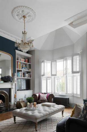 Maison Pippa Jones: salon avec grande baie vitrée, volets blancs, murs blancs et mur caractéristique bleu foncé, table basse ottomane boutonnée beige et tapis rose et blanc