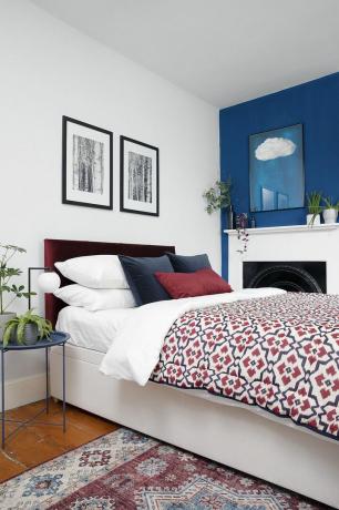 सफ़ेद दीवारों वाला शयनकक्ष, एक नीली फ़ीचर दीवार, बरगंडी मखमली हेडबोर्ड और लाल और नीले पैटर्न वाला बिस्तर, लाल फ़ारसी शैली का गलीचा और फ़्रेमयुक्त कलाकृति