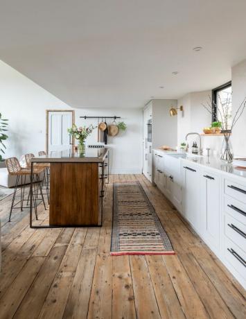 Piso de tábuas de madeira em plano aberto, cozinha rústica moderna com armários brancos, ilha de madeira escura e bancos de bar de tecido.