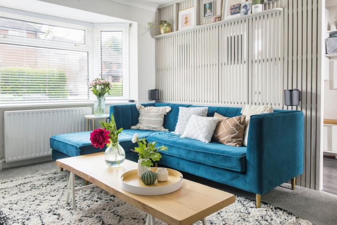 Sala de estar com paredes brancas, carpete cinza, tapete boho, sofá de veludo azul, mesa de centro de madeira e divisória de ripas cinza no corredor