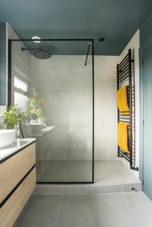 Kylpyhuoneessa harmaat seinä- ja lattialaatat, siniseksi maalattu katto, suihkukaappi mustalla kehyksellä ja musta patteri