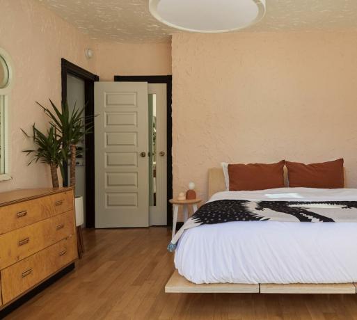 Dormitor vopsit în blush de coral cu pat jos din lemn, bufet retro, lemn negru, podea din lemn de esență tare, așternut alb, plantă