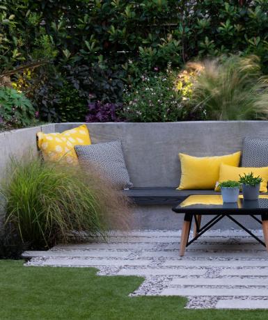אזור חצר אחורית מכוסה עם ספה חיצונית אפורה, כריות צהובות ואדניות גדולות מלאות בדשא