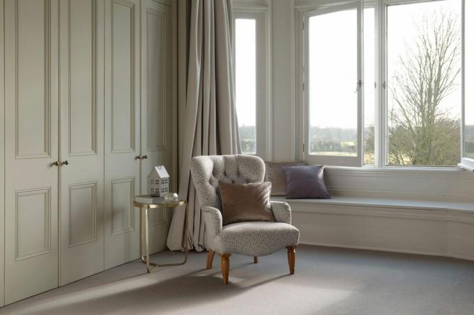 Neutral gepolsterter Stuhl im Schlafzimmer mit Teppichboden, hellgrünen Einbauschranktüren, Erkerfenster und vergoldetem Beistelltisch.