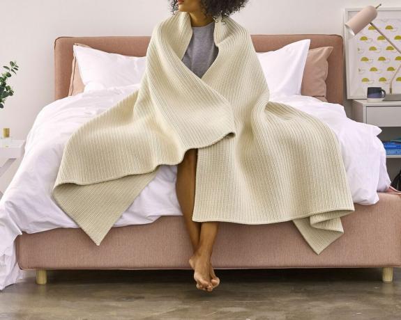 Una coperta a coste di cotone color crema avvolta intorno alle spalle di una donna seduta su un letto imbottito