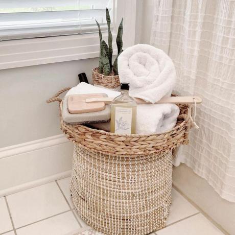 Maalaistyylinen pamperspa-kori, jossa on rullatut pyyhkeet, pesusieni ja huonekasvi