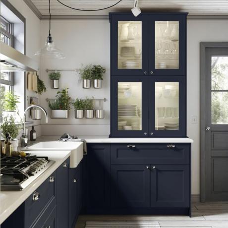 modrá kuchyně se skleněným přívěskem nad dřezem, bylinky zavěšené na stěně, světlé dřevěné podlahy, prosklená skříňka
