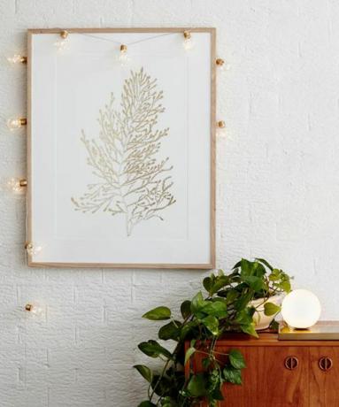 Medeninaste baterijske mikro festonske luči Lights4Fun so obešene okoli stenske umetnine z leseno omarico in sobno rastlino