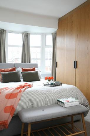 Ložnice s šedou postelí a lavicí, dřevěné vestavěné skříně, pruhované bílé a šedé povlečení a korálové doplňky