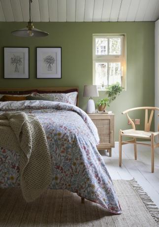 zielona sypialnia w stylu wiejskiego domu z kwiecistą pościelą, dziełami sztuki, drewnianym krzesłem, stolikiem, białymi deskami podłogowymi, sufitem shiplap i zawieszką wznoszącą się i opadającą