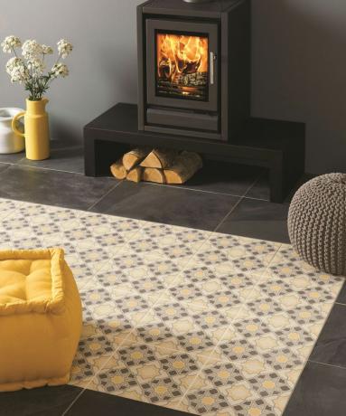 Šiltų pilkų ir geltonų atspalvių tradicinės raštuotos grindų plytelės su kilimėlio efektu, priešais riaumojančią ugnį.