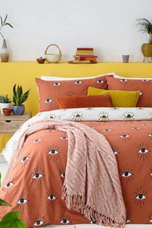 แต่งตาด้วยผ้าปูที่นอนสีชมพูนวลบนเตียงที่มีผนังสีเหลืองด้านหลังและต้นกระบองเพชรด้านข้าง 