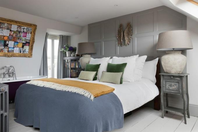 Dům Pippa Jones: hlavní ložnice s bíle natřenými podlahovými prkny, šedá obložená stěna, postel s modrými a žlutými hody a nouzové noční stolky s nadměrnými lampami