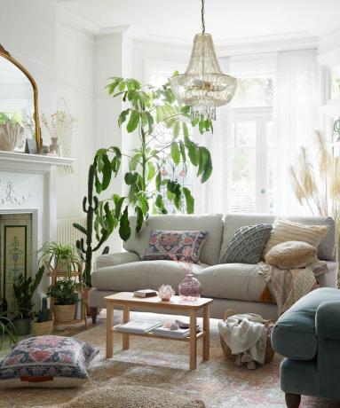 Boho obývacia izba s vrstvenými textíliami, hmatovými a vzorovanými rozptylovými vankúšmi na neutrálnej pohodlnej pohovke, korálkovým boho stropným príveskom, kaktusmi a veľkou rastlinou v kvetináči.