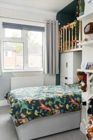 Cameră pentru copii cu pat acoperit cu așternut de dinozauri ascuns într-un alcov cu bârlog, accesibil printr-un perete de cățărat, deasupra – vopsit într-o culoare verde închis, cu balustrade din lemn