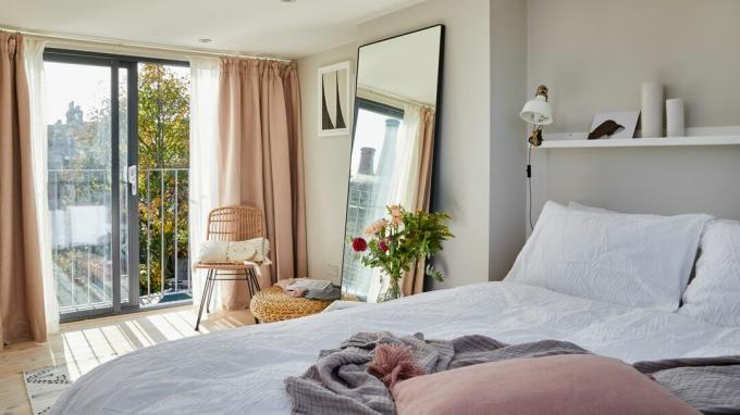 Tende rosa su porte scorrevoli in camera da letto con biancheria da letto bianca, specchio a figura intera, sedia in rattan e vegetazione all'interno