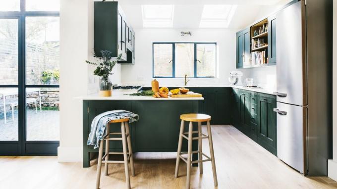 Een kleine keuken geschilderd in donkergroen en wit in een open leefruimte met een ontbijtbar, krukken en schiereiland