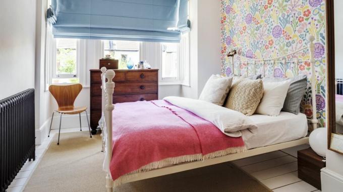 ห้องนอนพร้อมวอลเปเปอร์ลายดอกไม้ มู่ลี่ผ้าลินินสีน้ำเงิน ผ้าปูที่นอนสีชมพู และลิ้นชักโบราณ