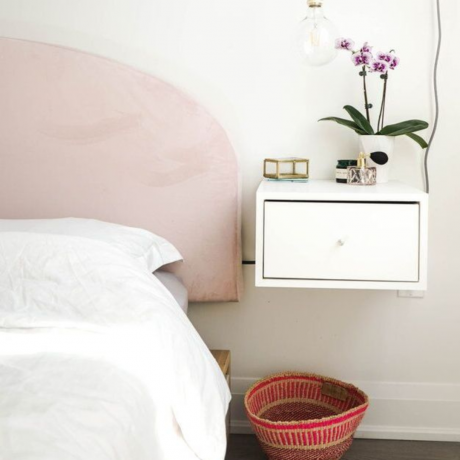 Lebdeča nočna omarica s predalom v beli barvi, okrašena z orhidejami v lončkih in drobnarijami, poleg ukrivljenega vzglavja v bledo rožnati barvi in ​​izpostavljenim obeskom v obliki žarnice, ki visi zgoraj.