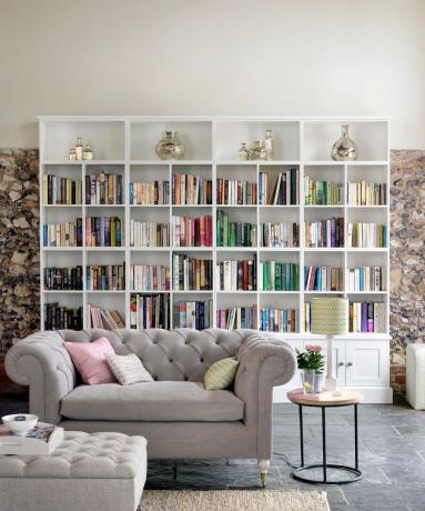 velké bílé police na knihy plné knih proti zdi z přírodního kamene s pohovkou chesterfield v béžové barvě