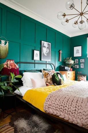 Pareti con pannelli verdi in una camera da letto principale con un letto di metallo, plaid gialle e rosa e cuscini fantasia