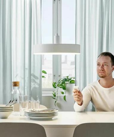 un uomo che attenua la lampada a sospensione NYMANE Ikea usando un telecomando, mentre è seduto al tavolo da pranzo