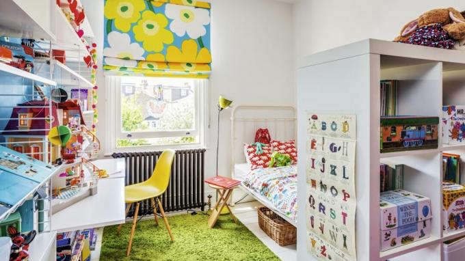 ห้องนอนเด็กสีสันสดใสพร้อมมู่ลี่ดอกไม้สีน้ำเงินและสีเหลือง เตียงโลหะสีขาว และหม้อน้ำแบบรีเคลม
