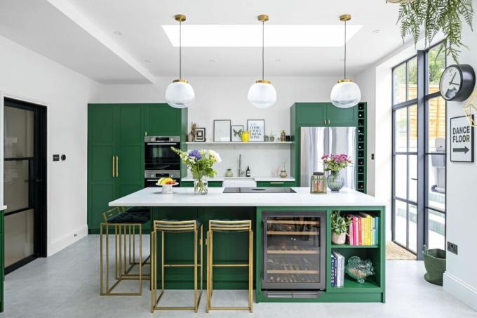 Žalia moderni virtuvė su sala; balto kvarco stalviršis, žalvarinės baro kėdės, žalvario ir stiklo pakabinami šviestuvai ir poliruotos betoninės grindys