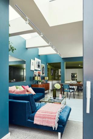 Гостиная с синим бархатным диваном, покрывалом и красочными подушками. Бетонный пол с ковриком и стеклянным журнальным столиком