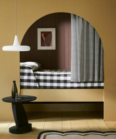การออกแบบห้องนอนซุ้มสีเหลืองพร้อมโต๊ะสีดำ โคมไฟเพดานสีขาว และผ้าปูที่นอนลายตารางโดย Little Greene Paint Company