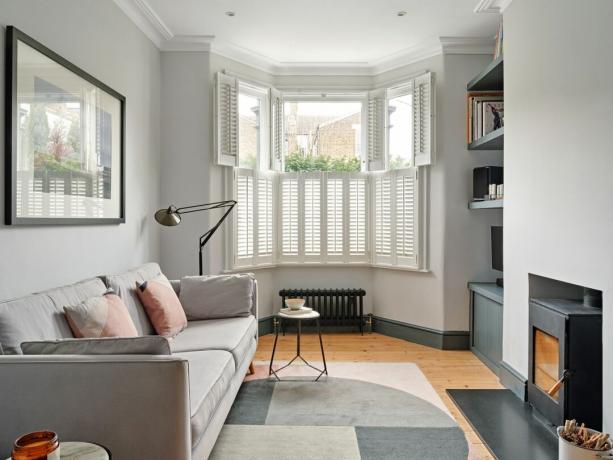 Biela obývačka s pecou na drevo, svetlosivou pohovkou, šedým geometrickým kobercom a bielymi okenicami v arkierovom okne