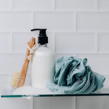 Primer plano de productos de ducha, cepillo de baño y lufa sobre azulejos blancos del metro.