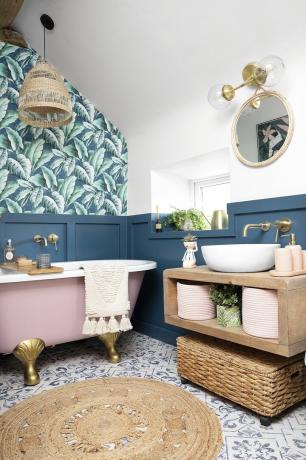 Kamar mandi berpanel biru dengan wallpaper palem, lantai ubin berpola, permadani rami, dan unit meja rias apung yang digantung di dinding pedesaan