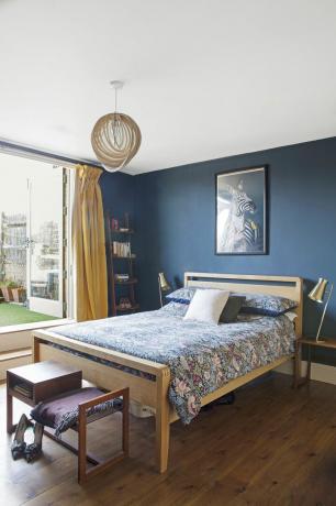 Makuuhuoneessa tummansiniseksi maalattu seinä, puulattia, tammi sängyn runko ja kukkaiset lakanat rakenteellisella tammiriipusella