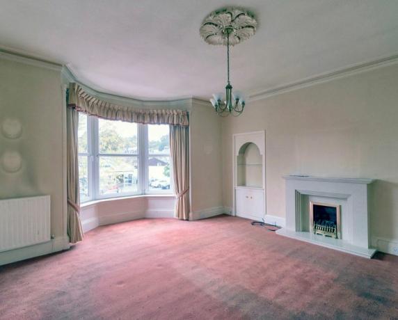„Vorher“-Aufnahme zeigt Wohnzimmer mit cremefarbenen Wänden, Kronleuchter und rot-rosa Teppich